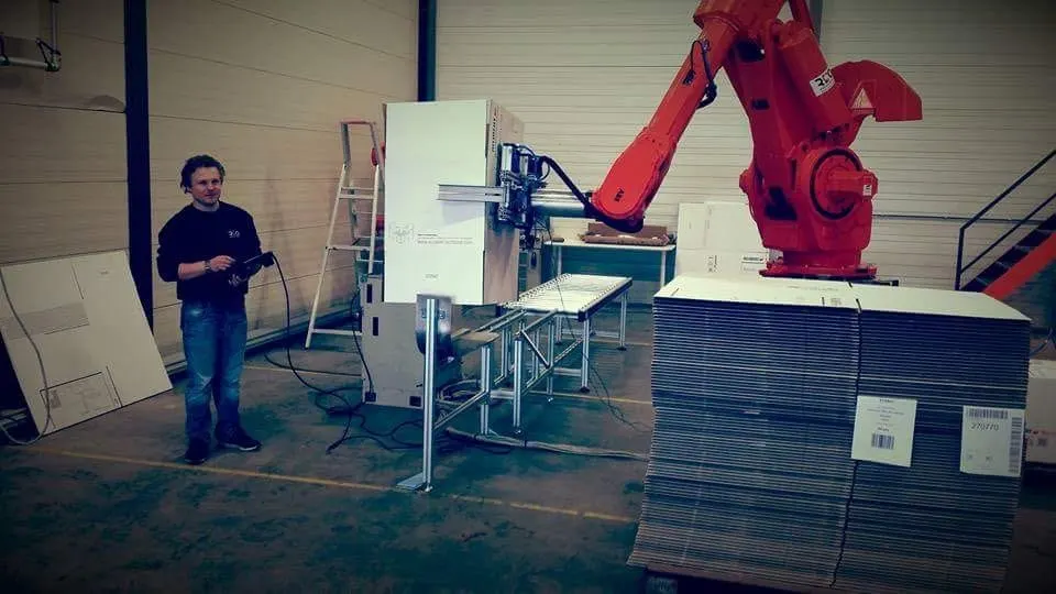 Mike van De Machinedokters programmeert een ABB-robot die een doos uitvouwt, naast een stapel platte doosuitslagen.