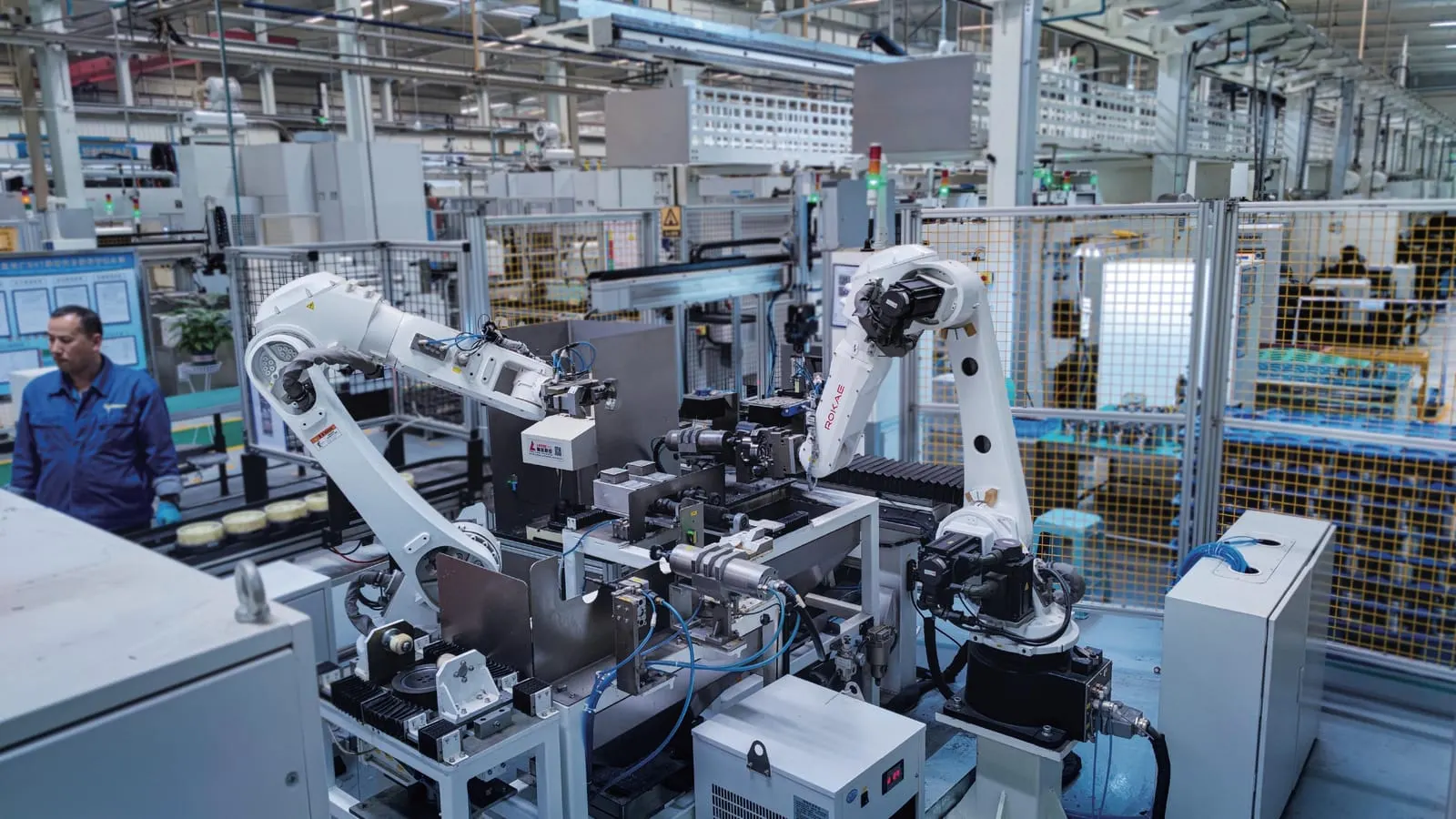 Twee industriële robots in een afgeschermde cel, met een operator buiten het hekwerk.